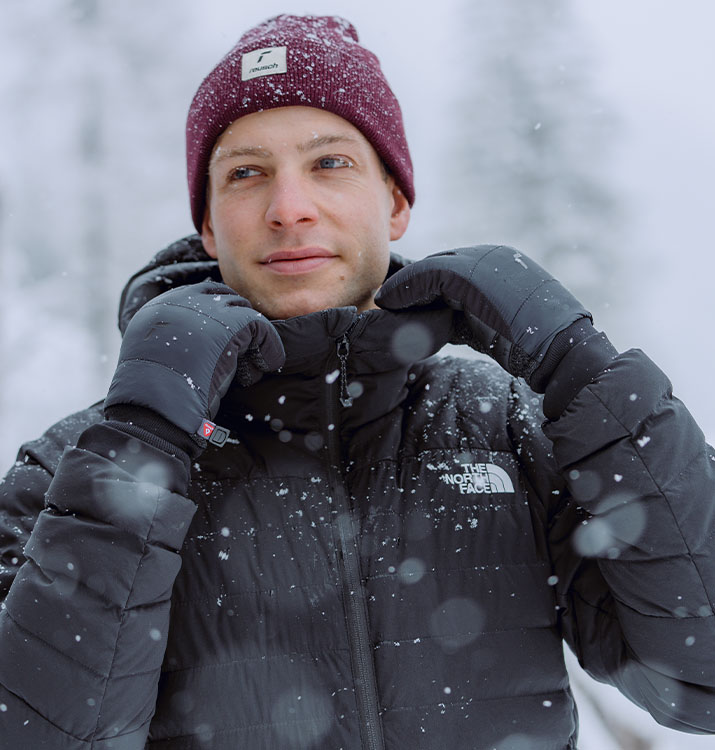 Mann im schwarzen Daunenjacke und burgunderroter Mütze zieht den Reißverschluss hoch, während er im Schneefall schwarze Reusch-Winterhandschuhe trägt.