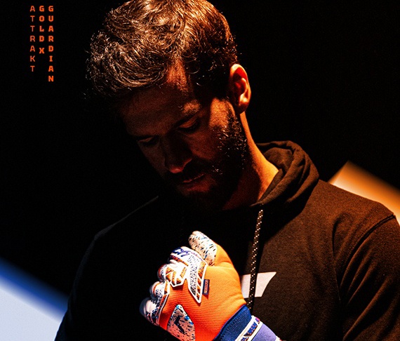 Torwart Alisson Becker im schwarzen Hoodie, der seine auffälligen, orange-blau-weißen Reusch Gold X Guardian Torwarthandschuhe anzieht, im dramatischen Licht.